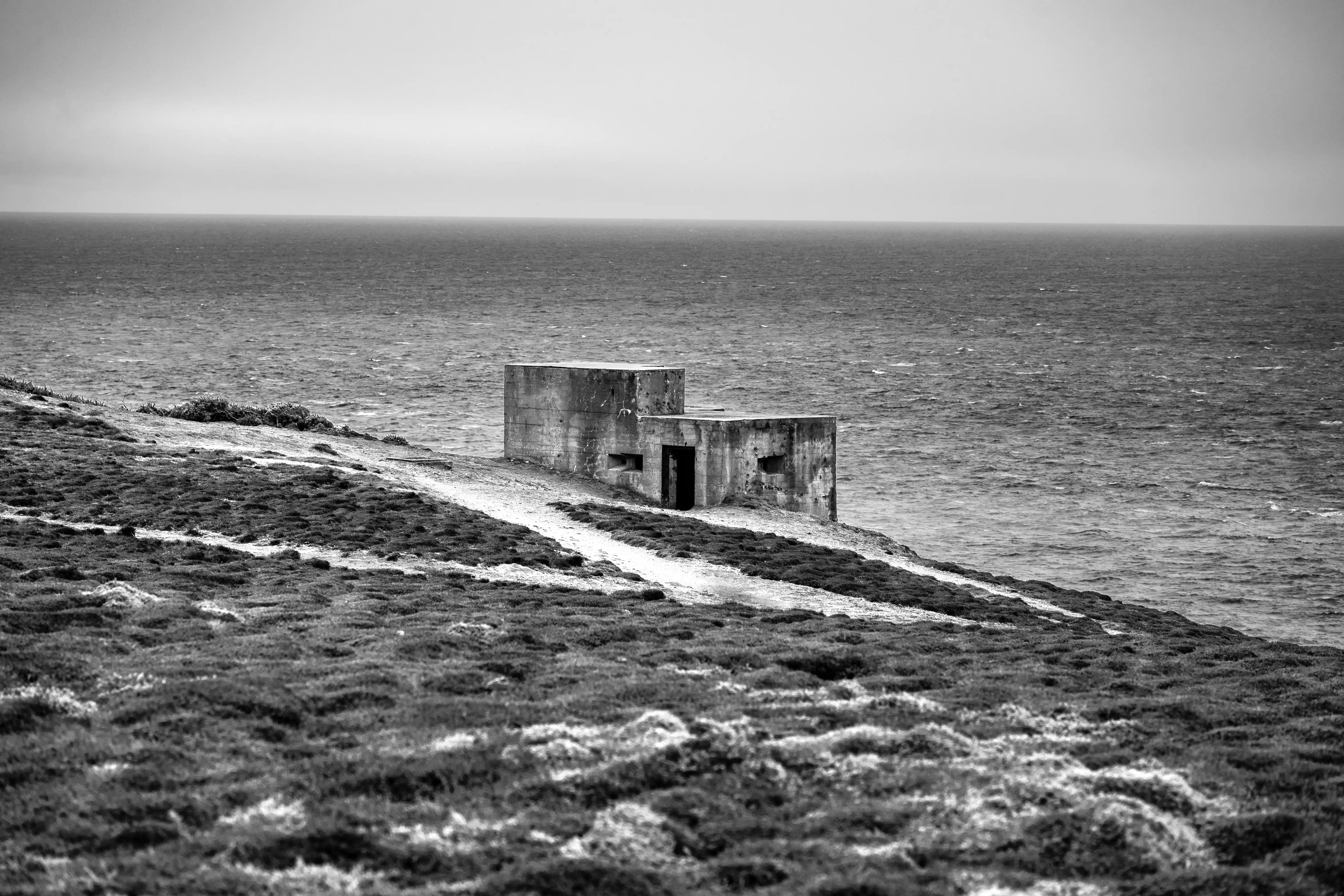 A black and white image of an old concrete military bunker on a windswept grassy cliff next to a dark, choppy sea.